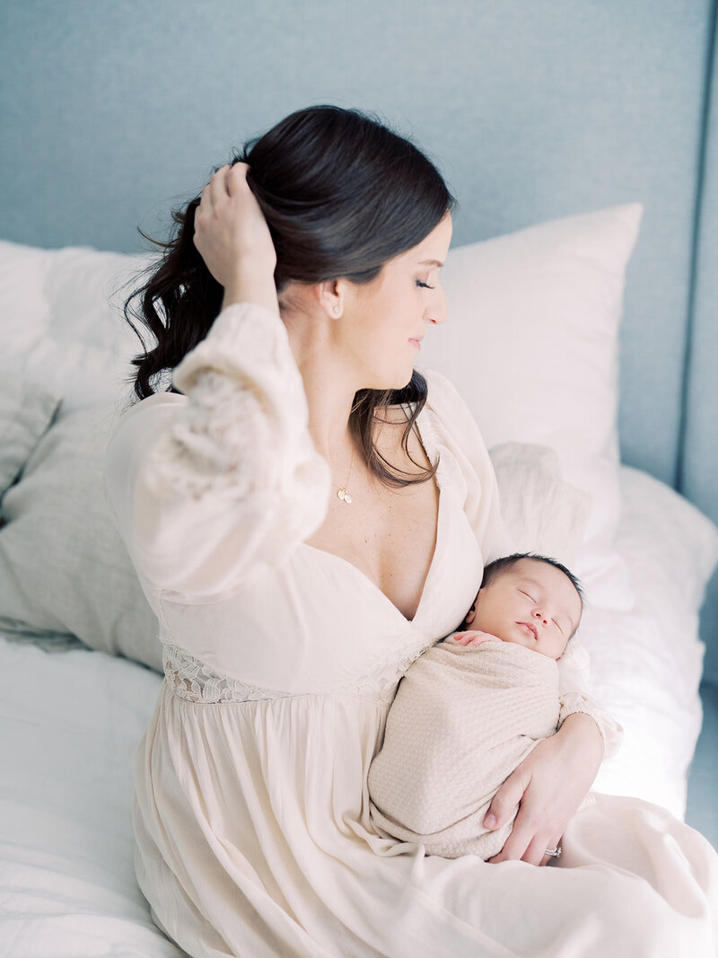 Mother with brown hair holds newborn baby as she sits on edge of bed with one hand up in her hair.