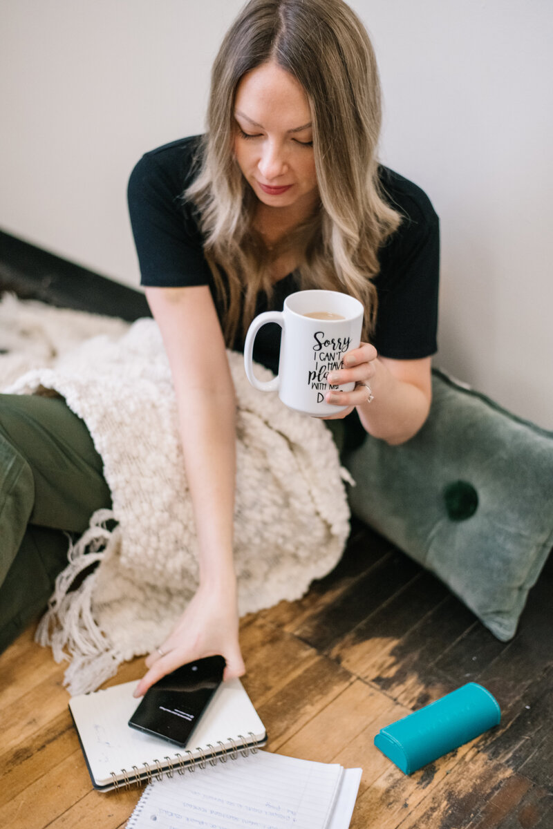 Sammy sitting on the floor with a blanket holding a mug and her phone
