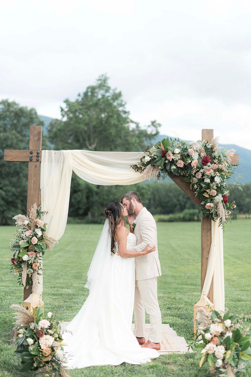 New bride and groom sealing vows with a kiss beneath a wooden pergola draped with white linen and floral arangements.
