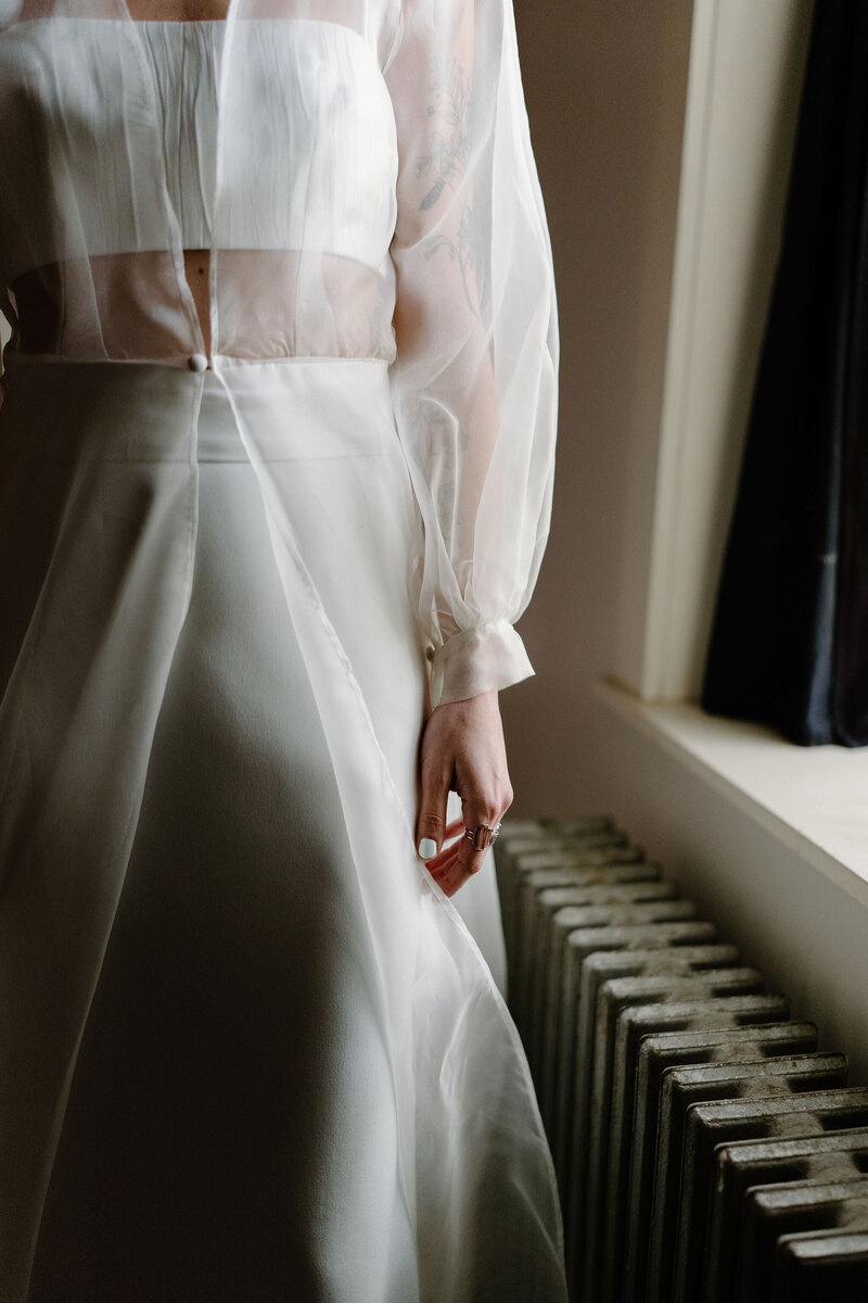 Close-up photograph of bride (long sleeve sheer wedding dress) standing by a window