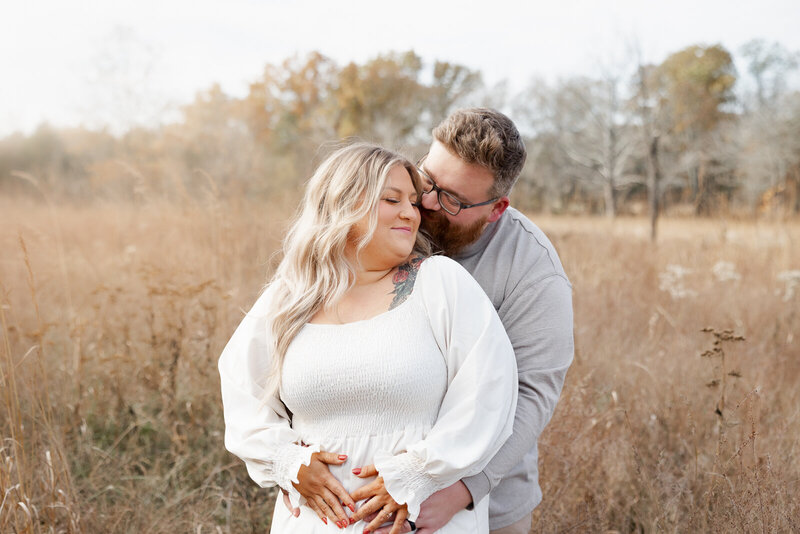 Expecting couple standing and embracing in a field during soft morning light.