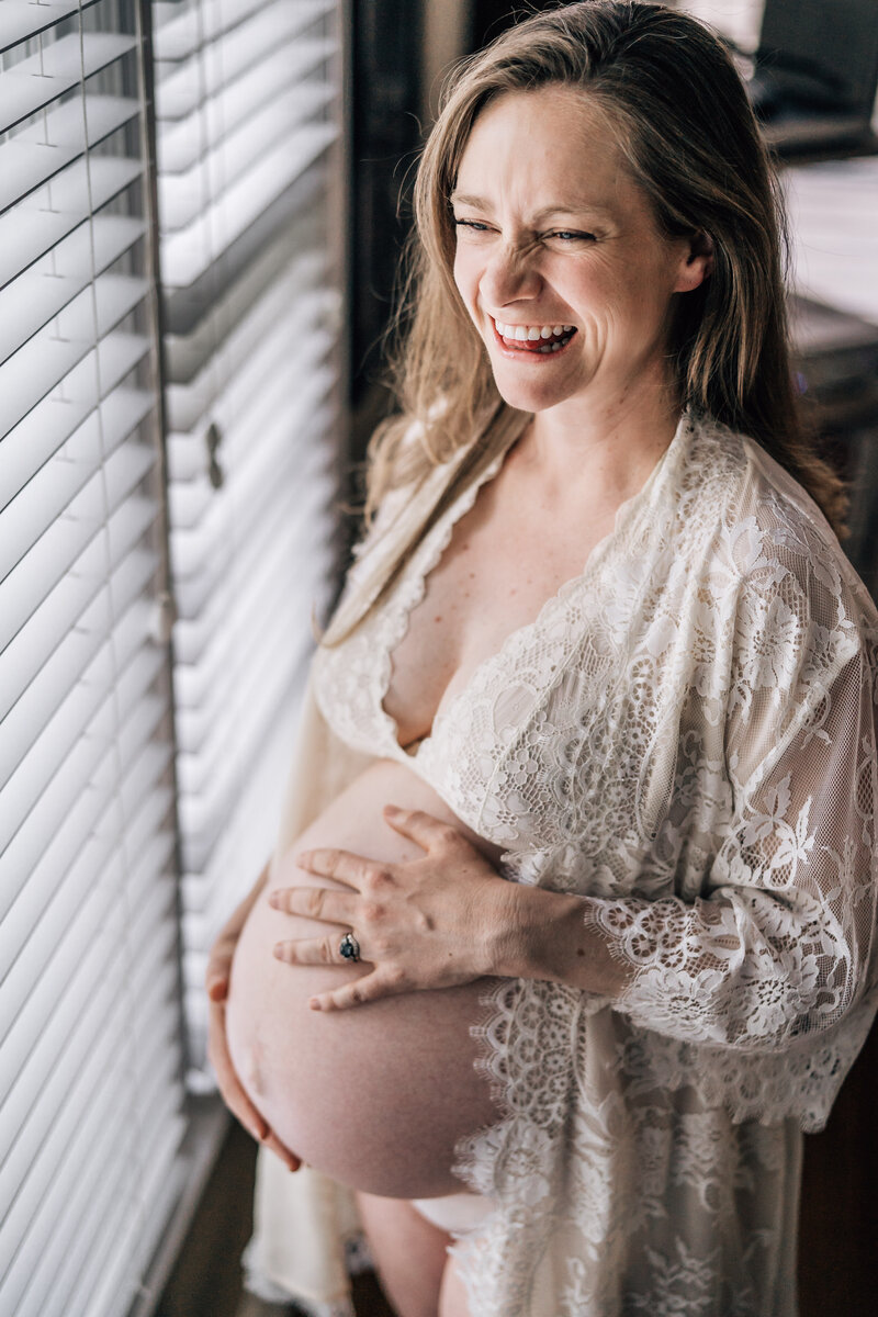 A pregnant woman standing by a window, dressed in a lace robe, smiling joyfully as she cradles her bare belly, bathed in soft, natural light.