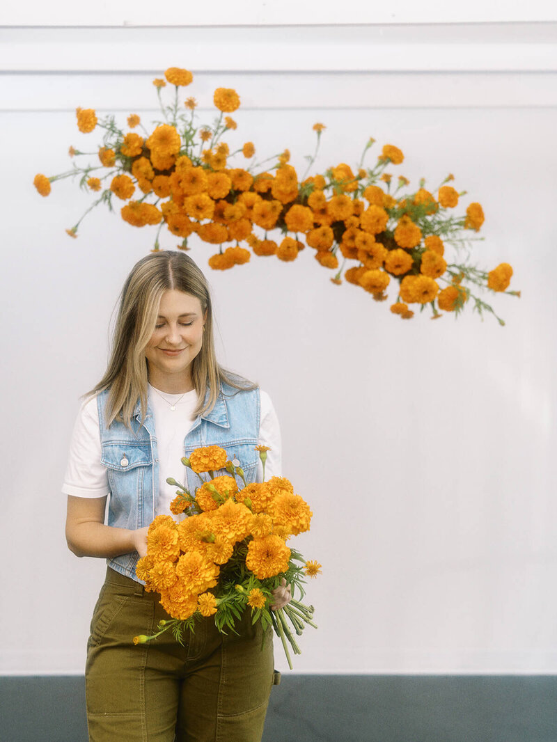Oregon wedding florist portrait. Florist is holding a bundle of orange marigolds. Above and behind her, there is a suspended floral installation of all orange marigolds. The background is a white wall. Setting is Eugene, Oregon.
