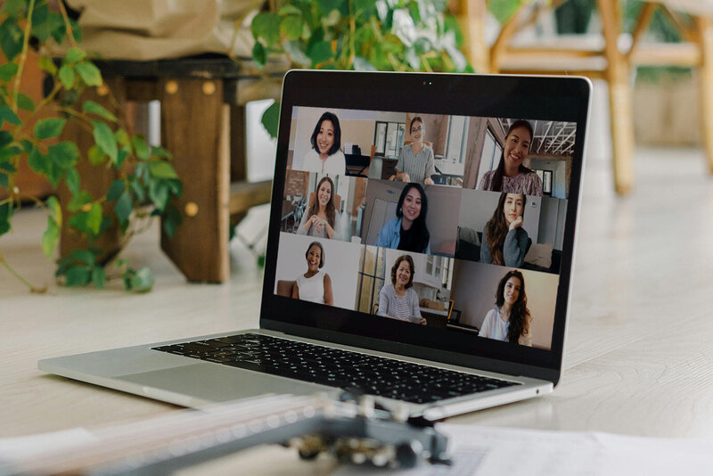 Laptop displaying a video call with nine smiling participants in a cozy indoor setting. Nearby, a guitar and papers rest on the floor. Vibrant, collaborative atmosphere.