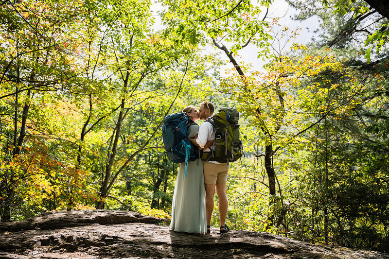 A couple wearing hiking backpacks stand on a rock with forest in the background and give each other a kiss. They're photographed during a brief pause on their hike to Dark Hallow Falls in Shenandoah National Park.