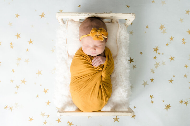 baby girl wrapped in yellow sleeping in tiny bed on backdrop with gold stars