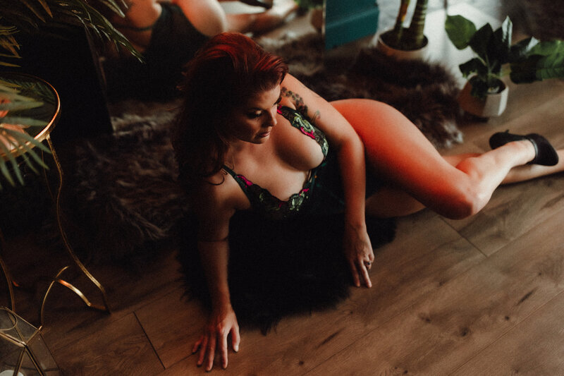 Woman with long red hair lays on rug under warm lights in Tampa boudoir studio, wearing black mesh lingerie
