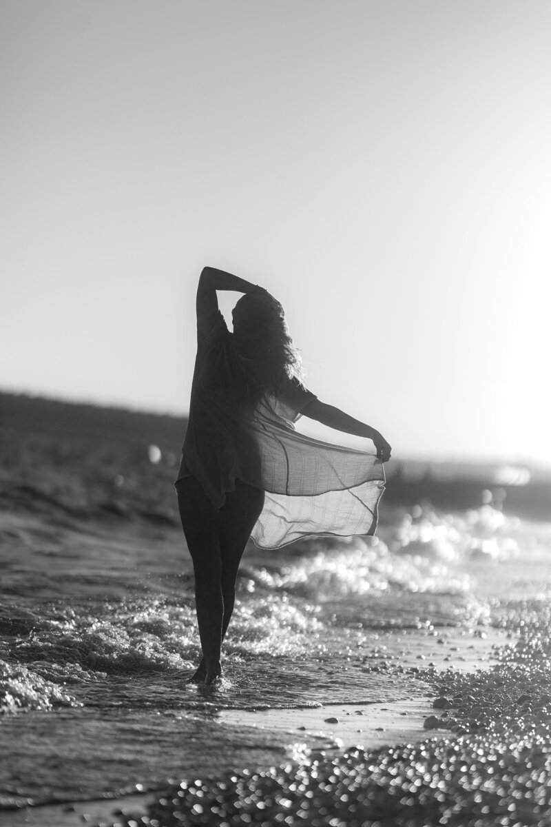 Black and white photo of silhouette of a women walking along the shoreline with one arm stretched overhead and the other holding out the fabric of her sheer dress.
