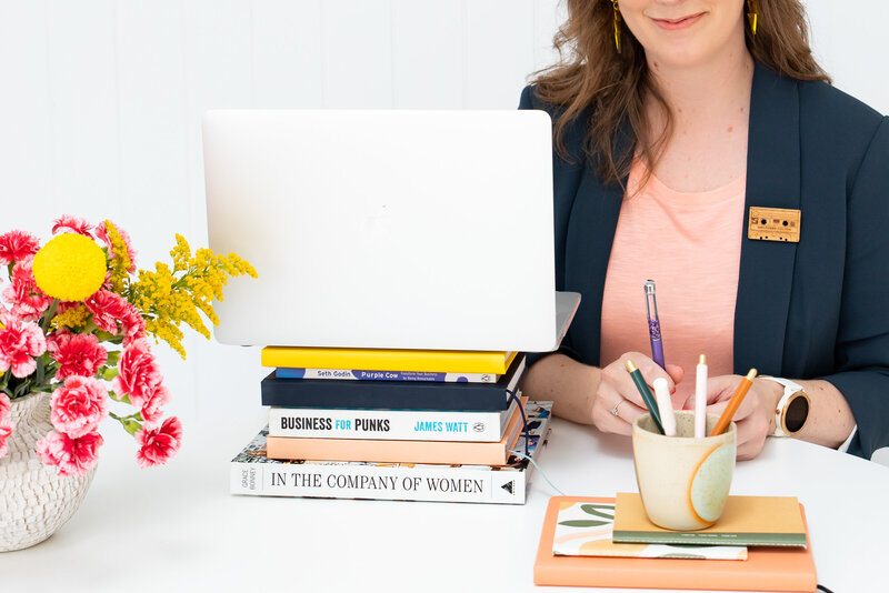 Melissa-Packham_A-Brand-Is-Not-A-Logo_Melissa-face-out-of-shot-wearing-navy-blazer-with-wooden-cassette-brooch_sitting-at-table-making-notes_notebooks-pen-holder-laptop-atop-pile-of-books-floral-arrangement_Landscape