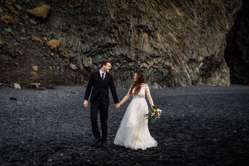 A bride and groom walk on a black sand beach at their Southern Iceland elopement.