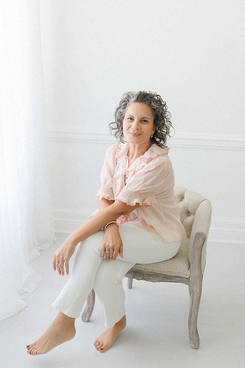 Nestled against a plain white backdrop, a woman with curly hair sits barefoot on a small chair, donning a light pink blouse and white pants. This serene scene captures the essence of simplicity beloved by Destin FL family photographers.