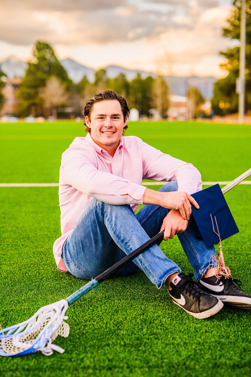 NAU senior guy male holding lacrosse stick and graduation cap on lawn on Northern Arizona University campus with peaks behind and sunset