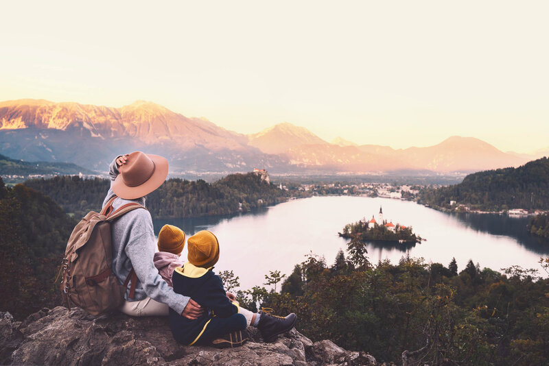 Person sitting with two children on a rocky viewpoint, overlooking a lake with a small island and surrounding mountains at sunset.