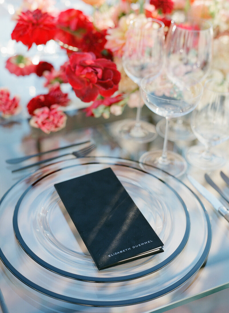 Wedding place setting design, glass plates with black rims, crystal wine glasses, and bright red carnations.