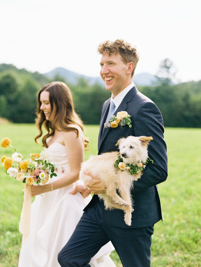 A smiling bride and groom carry the bouquet and puppy dog also adorned with flowers after the wedding ceremony at destination wedding venue Paint Rock Farm in North Carolina, by film photographer My Sun and Stars Co.