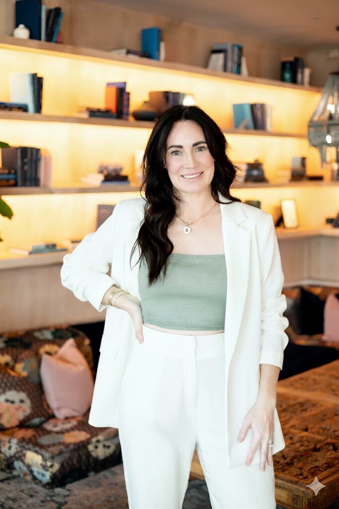 Woman with brunette hair in white suit with hand on hip in a room with books behind her.