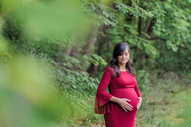 A pregnant woman in a red dress hugging her belly at a park.