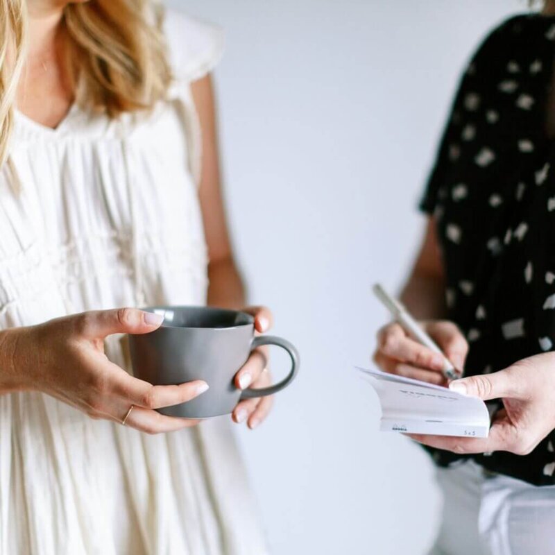 Two women standing together, one with a coffee mug in her hands and one writing on a notepad