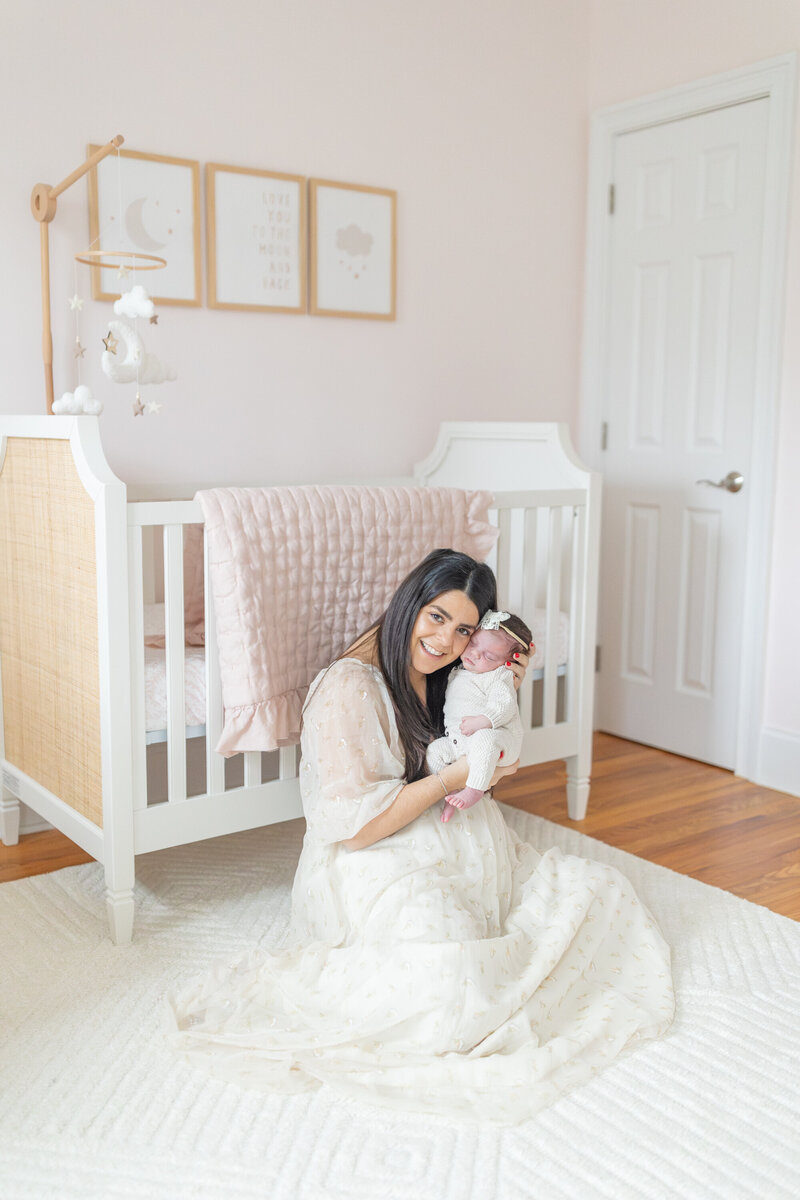 A mother sits on the floor of the nursery next to the crib, cradling her newborn in her arms. Soft natural light fills the room, creating a warm and intimate moment during an in-home lifestyle newborn session.