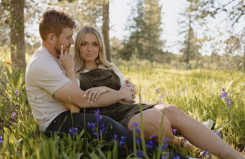 Man and woman holding hands in a field with dried grass and sagebrush. They're looking at each other and smiling.