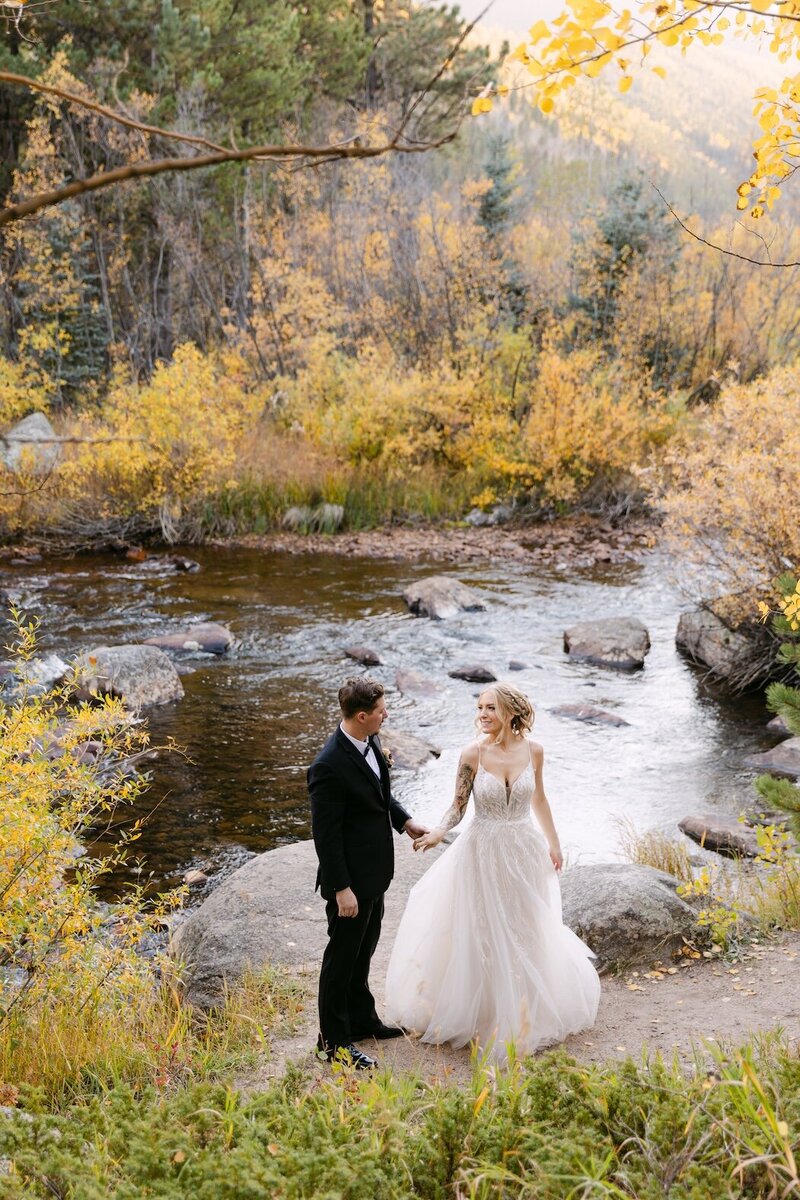 Bride and groom standing beside a mountain river surrounded by golden aspens at Wild Basin Lodge — fall Colorado mountain wedding portrait