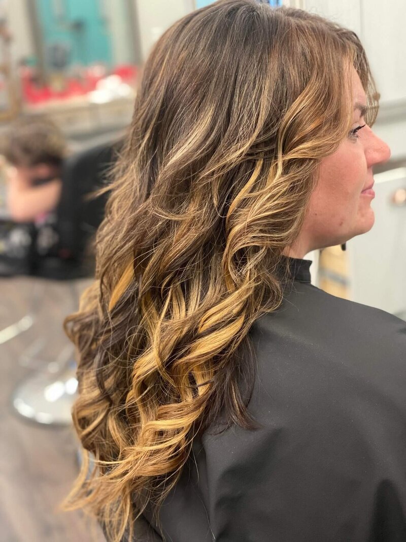 A woman with long, curly hair featuring golden highlights, seated in a salon chair and showcasing a glamorous hairstyle created by 212 Salon, Spa, & Barbershop.