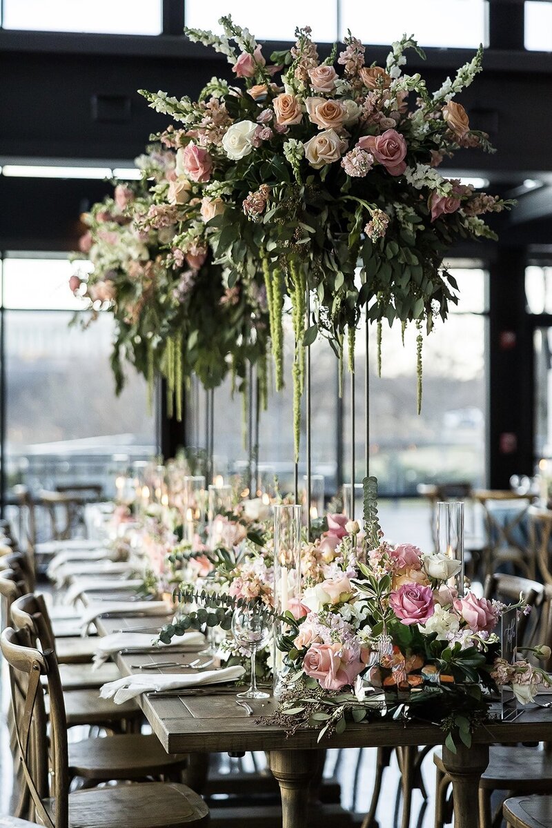 Elegant wedding reception table with cascading floral centerpieces of blush, peach, and ivory roses, white hydrangeas, and lush greenery, paired with wooden chairs and soft candlelight, designed by Wells Design Co.