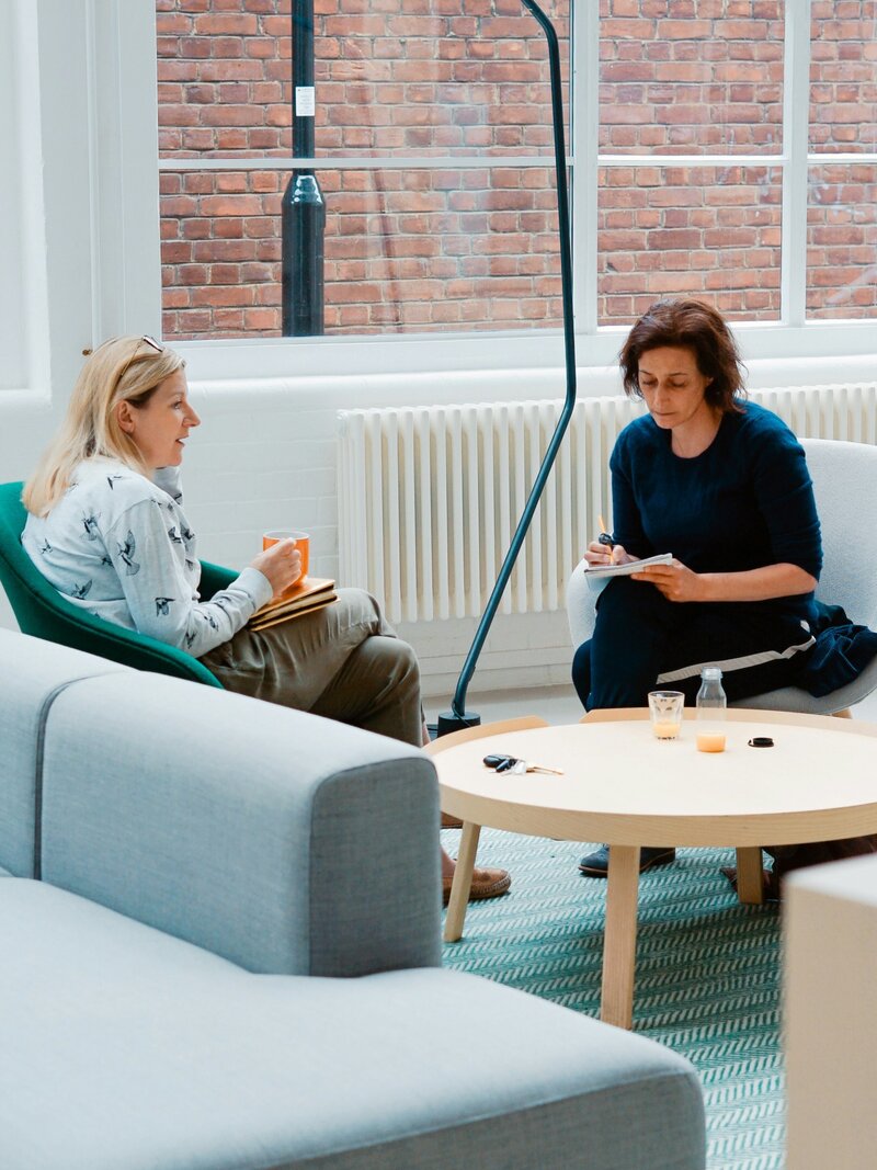 Two women sitting on chairs appearing as though one is talking with the other to help her.