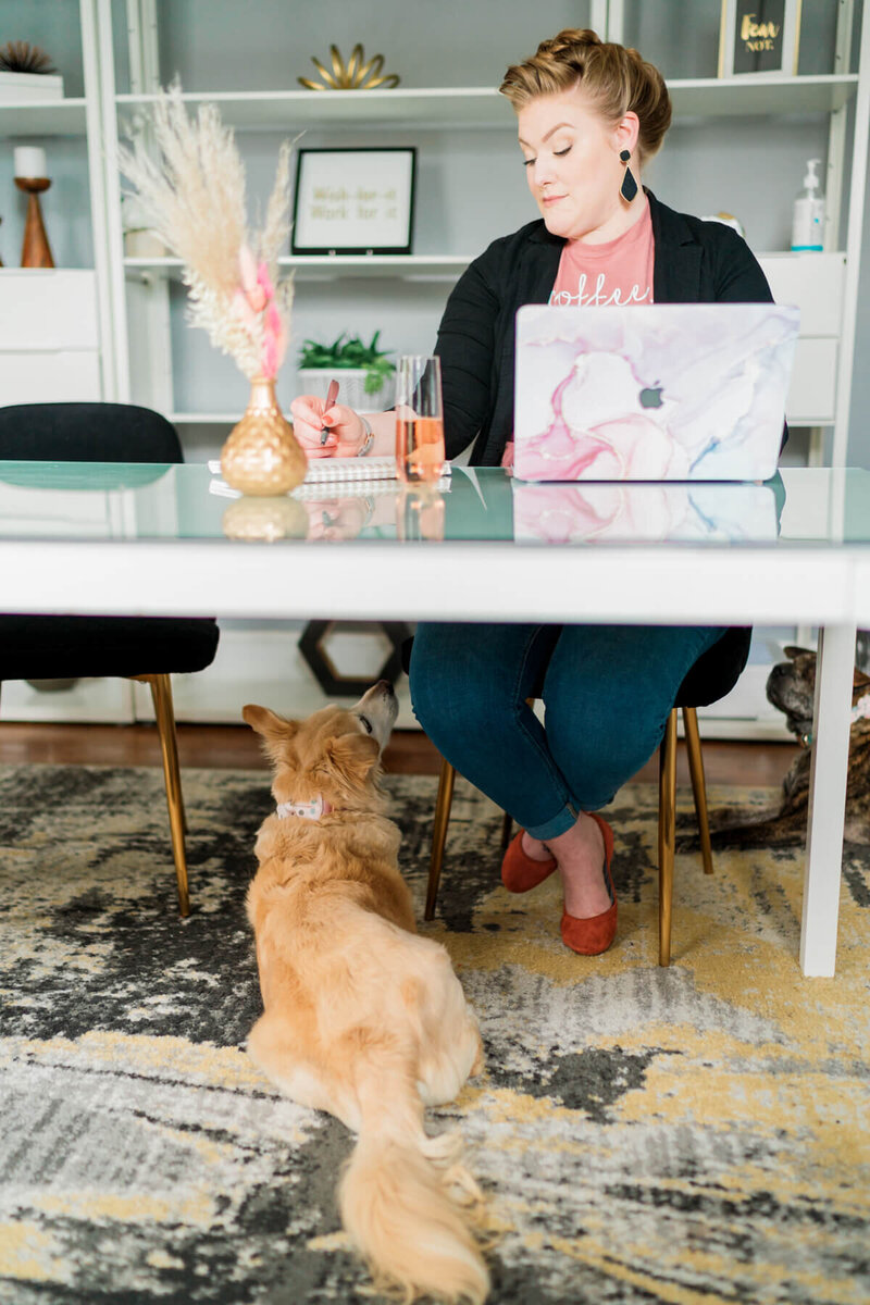 Becca Mai sitting at a table working while her dog Carly is laying at her feet