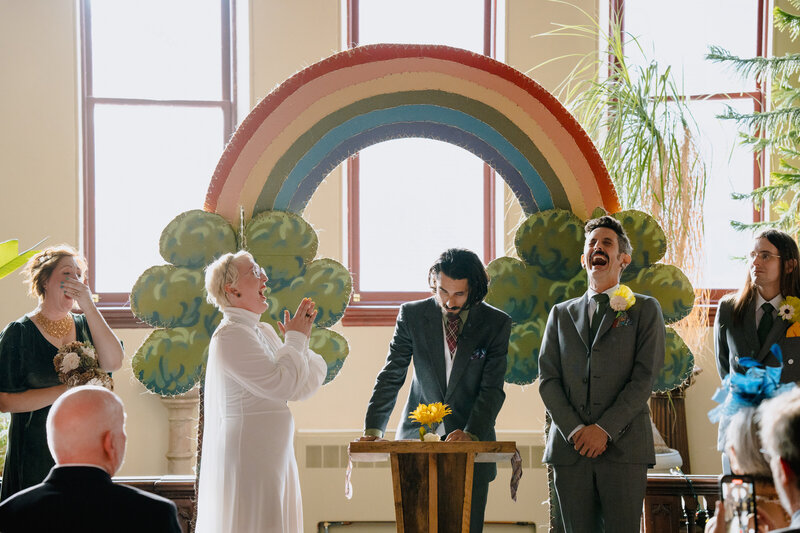 A couple standing up together during their wedding with the officiant and a rainbow behind them.