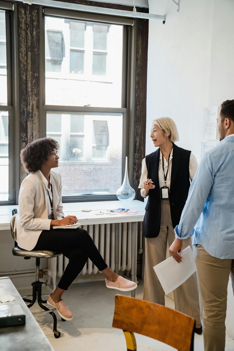 Three professionals collaborating in bright office space - woman with curly hair seated at desk, blonde woman standing presenting, and man in blue shirt listening, representing corporate team dynamics.