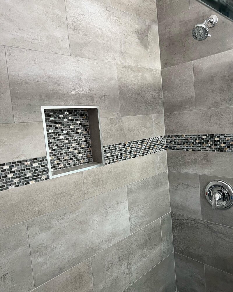 A newly remodeled shower with large grey tiles placed horizontally. There is chrome hardware and a black and white small tiled shampoo box on the left hand wall.