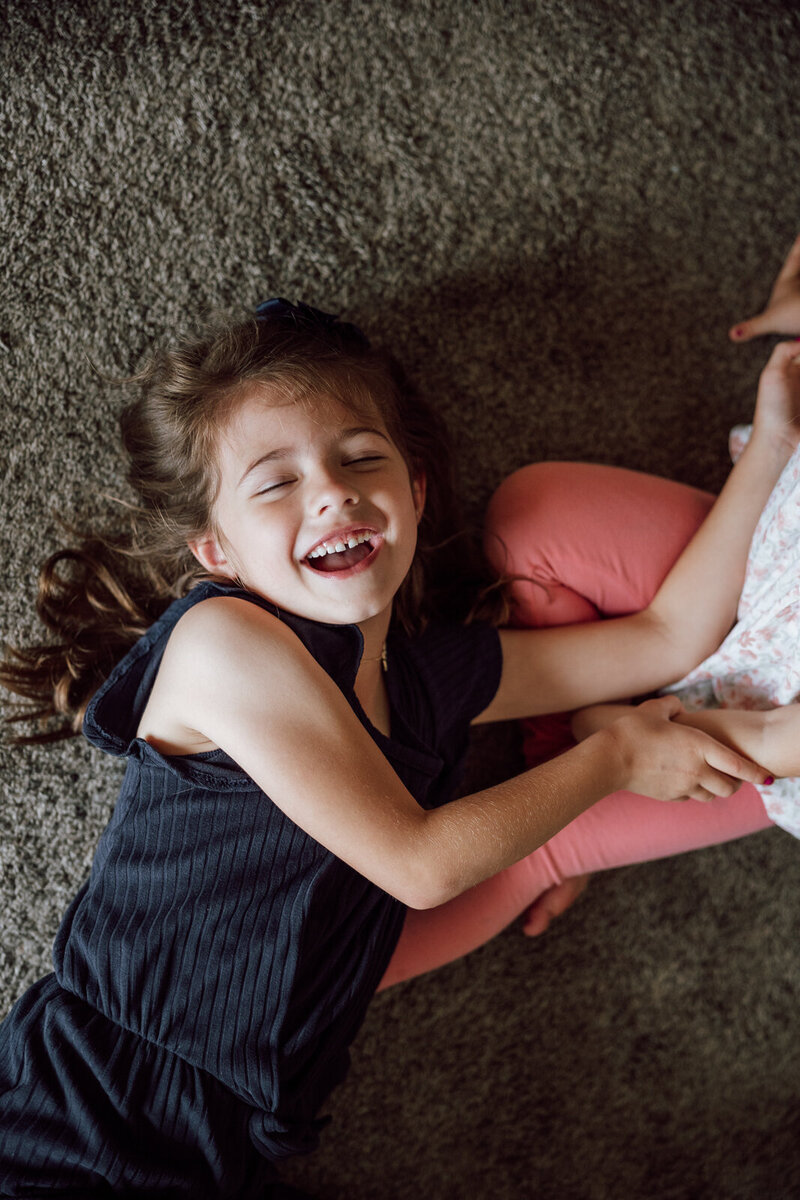 Little girl laying on the living room floor playing with sister and she's laughing. 