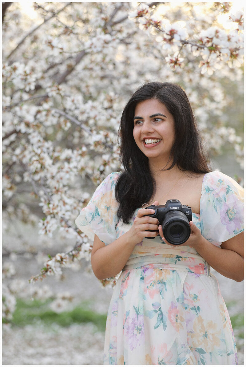 Female photographer holding a professional camera and gazing directly at viewer while posing in almond orchard with white blossoms during sunset