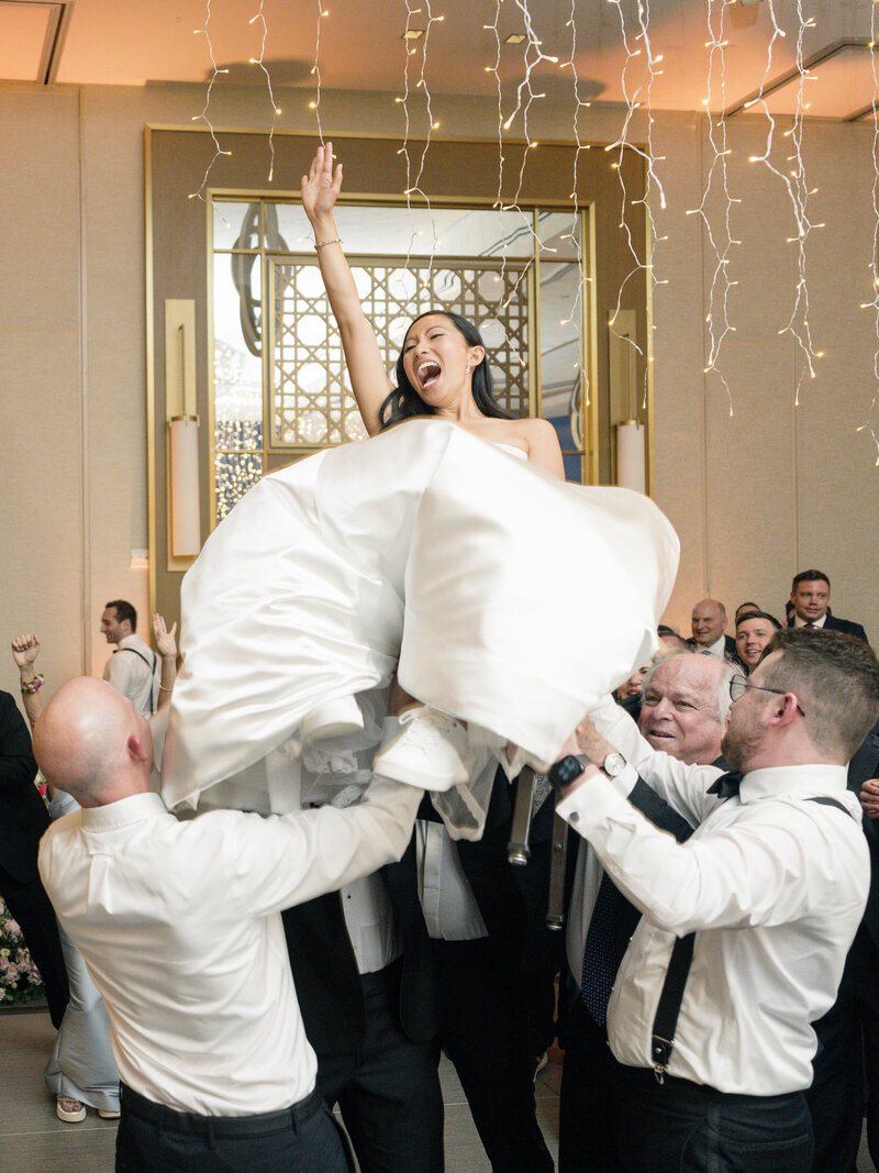 A joyous bride is lifted into the air by a group of people, surrounded by smiling guests. She is wearing a white dress and sneakers, while string lights hang from the ceiling, adding to the festive atmosphere.