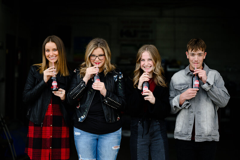 Senior photos in denver of three girls in black and red in an old garage laughing while  holding glass bottles of dr. pepper