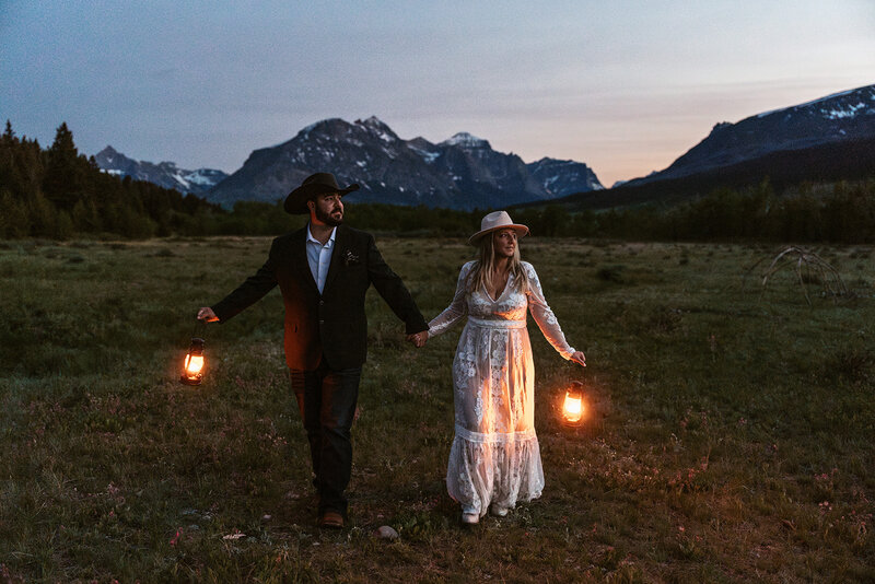 A couple walks hand in hand through an open field at sunset in Glacier National Park, holding lanterns that softly glow against the mountain backdrop, blending western charm with romantic adventure, captured by Sydney Breann Photography.