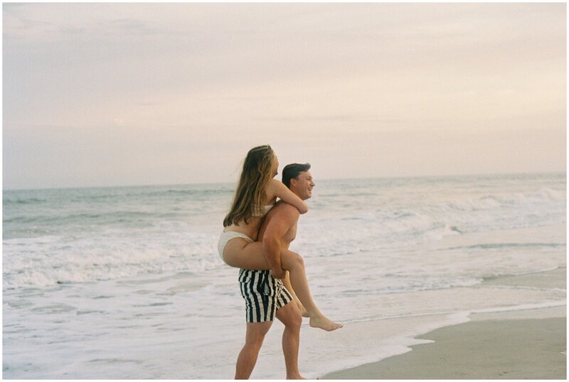 A man gives his fiancee a piggy back ride on the beach at sunset