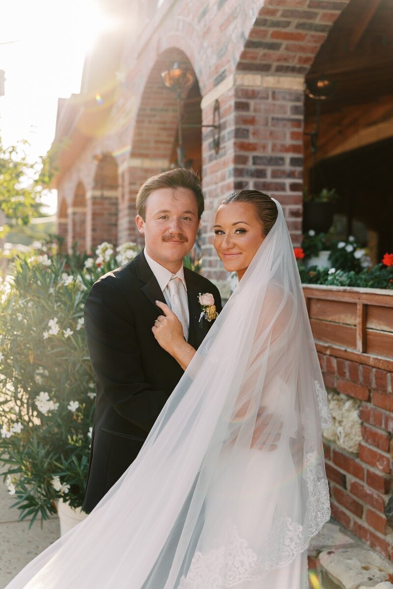 A bride and groom pose together outside by a brick wall with the bride’s veil flowing around them, at Grbic Events, a wedding venue in St. Louis.