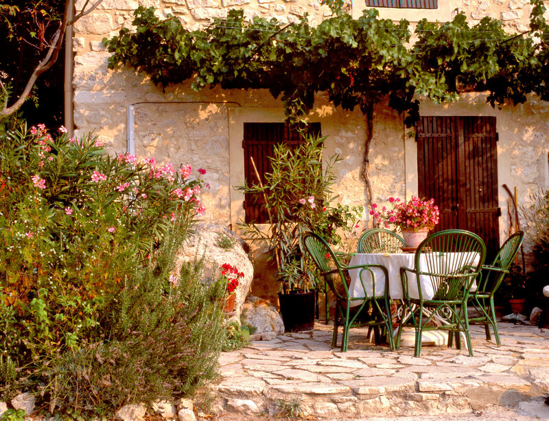 A rustic stone house with wooden shutters is adorned with lush green vines. In the foreground, a patio features a circular table with a white cloth surrounded by green chairs, amidst vibrant pink flowers and greenery.