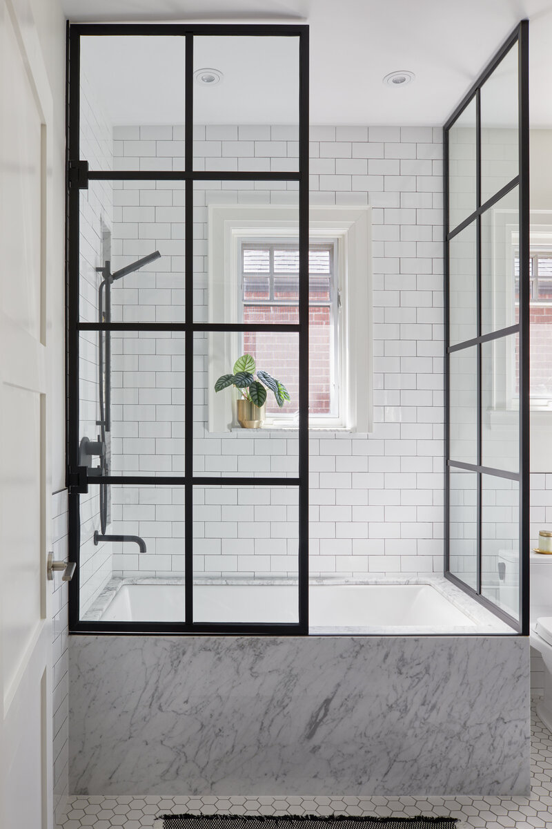 Side view of a rectangular marble bathtub surrounded by white subway wall tiles and a paned glass shower enclosure and a small window with a potted plant