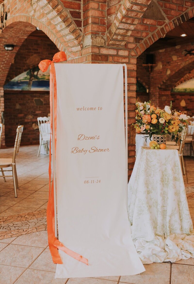 A decorated welcome sign for Desmi’s baby shower stands beside a floral arrangement, at Grbic Events, a wedding venue in St. Louis.
