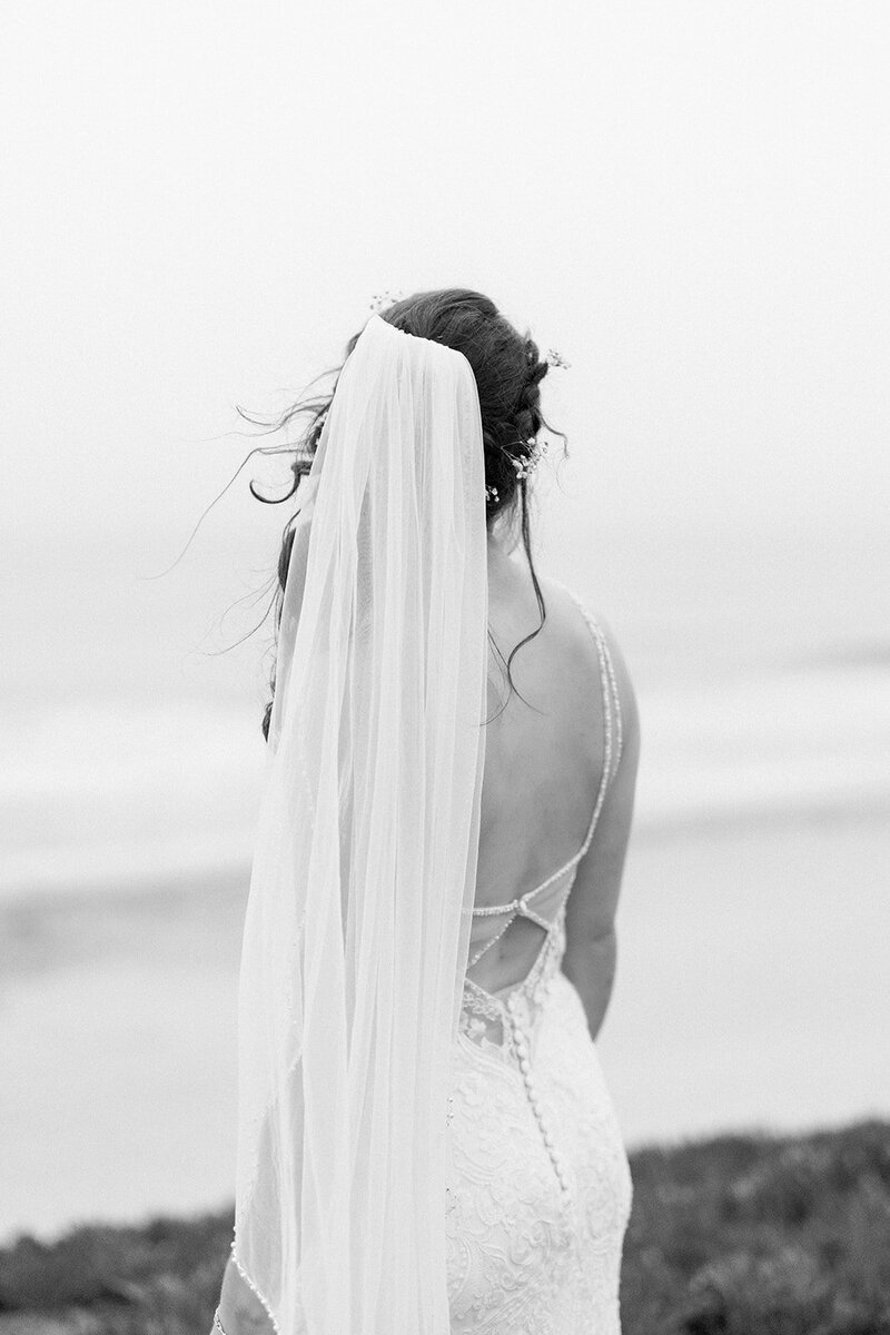 This black-and-white image captures a stunning California bride gazing out at the Pacific Ocean. Her veil flows softly in the breeze, creating a timeless and serene moment, perfect for a California wedding. The intricate lace detail of her gown contrasts beautifully against the backdrop of the ocean, making this photo an unforgettable representation of a coastal bridal portrait. Ideal for showcasing the romance and elegance of California weddings.