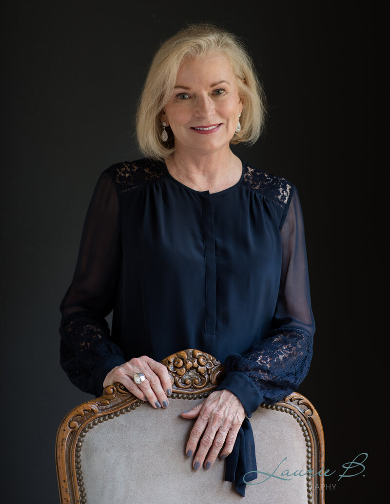 A portrait of  Karen McCall  in a blue dress, smiling and hands resting on the back of a chair