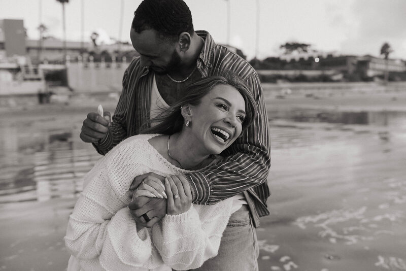 Black couple laughing and embracing during a joyful beach engagement session, captured in black and white with water and buildings in the background.