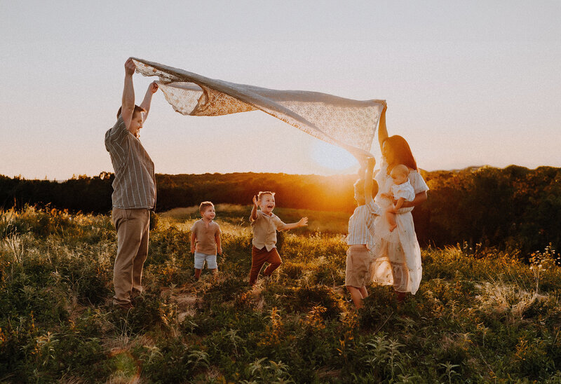 mother and father holding a quilt like a bridge for their young children to run under.