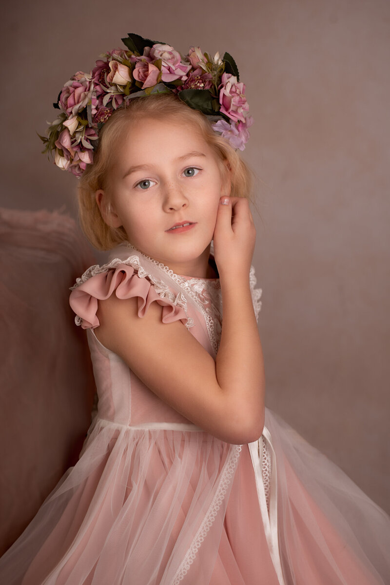 Delicate fine art portrait of a young girl wearing a soft pink dress and a floral crown, posed elegantly against a neutral backdrop. Captured in Overland Park, Kansas, this timeless and graceful setup is perfect for families seeking artistic and refined child photography in the Kansas City metro area