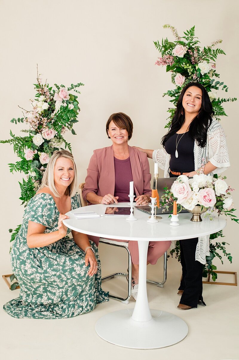 Group photo of 3 florists smiling at camera surrounding a table while working on laptop and surrounded by flowers