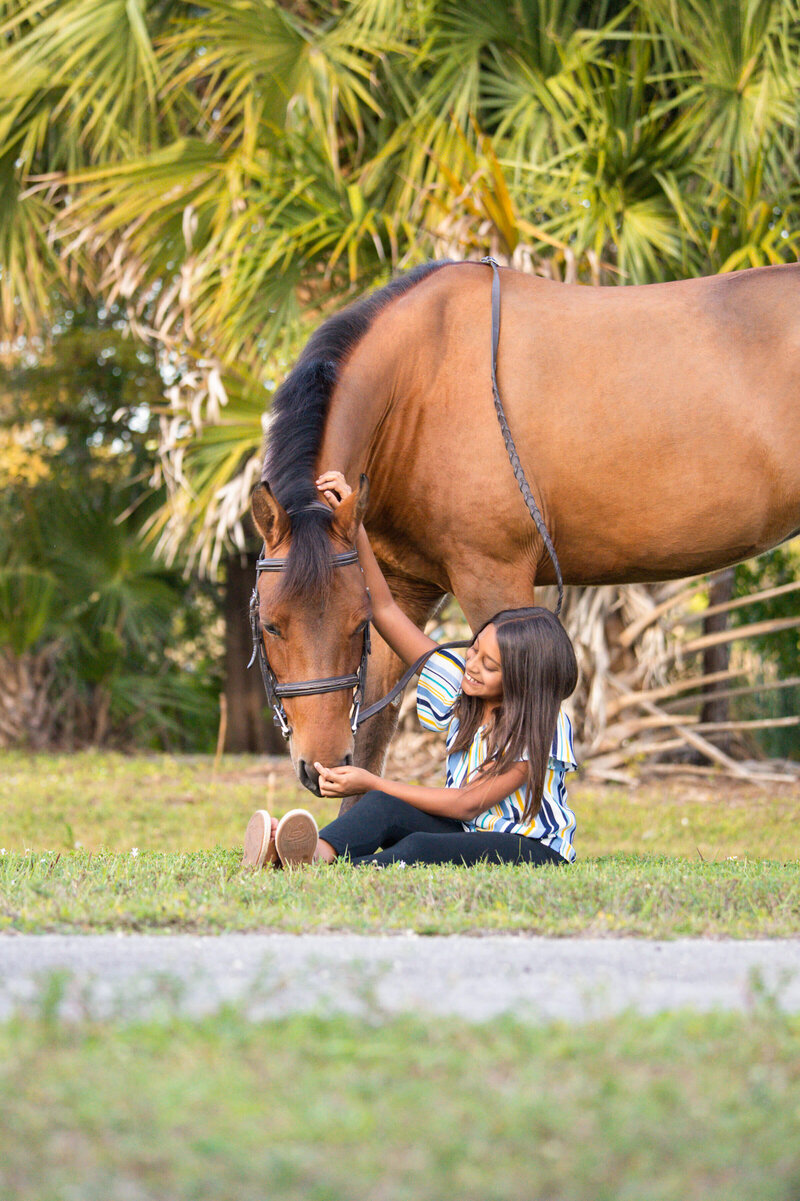 young girl sitting on the ground petting her horse's nose | south florida professional equine and pet photographer