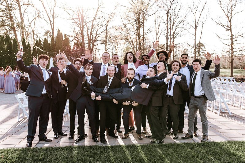 Group of joyful men, cheering with their arms up while horizontally holding groom, who is laughing and cheering with his arm up.